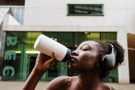 Portrait of young beautiful african woman in headphones drinking water with closed eyes outdoorsの写真素材