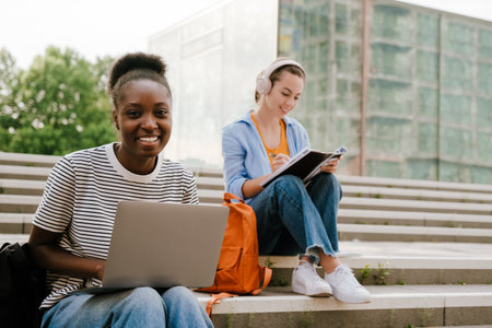 Young multiracial student women doing homework while sitting on stairs outdoorsの写真素材
