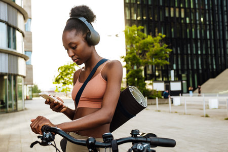 Young beautiful calm sporty african woman in headphones with yoga mat holding and looking on her phone, while standing on the street with bicycleの写真素材