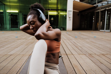 Young beautiful sporty african woman in headphones exercising on yoga mat lying near modern building...