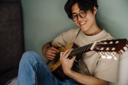 Young asian man in headphones playing guitar while sitting on bed at home