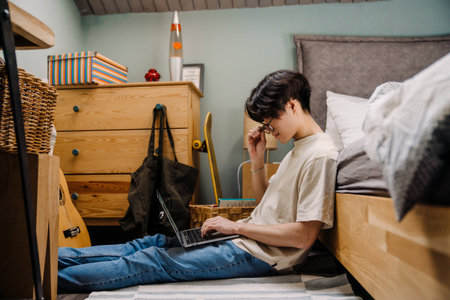 Asian teenage guy using laptop while sitting on floor at homeの写真素材