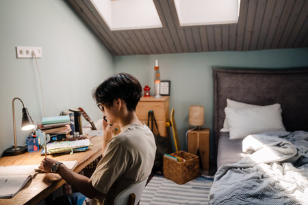 Asian teenage guy using cellphone while sitting at desk and studying at homeの写真素材