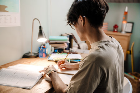 Serious asian teenage guy studying with book while sitting at desk at home