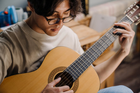 Young asian man in headphones playing guitar while sitting on chair at home