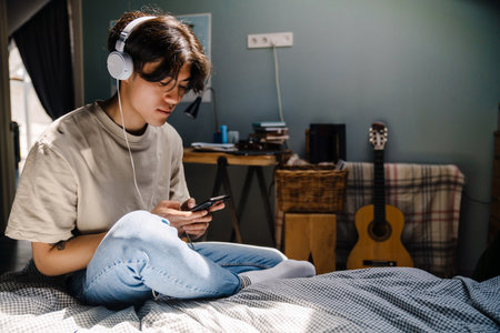 Young asian man in headphones listening music and using mobile phone at homeの写真素材