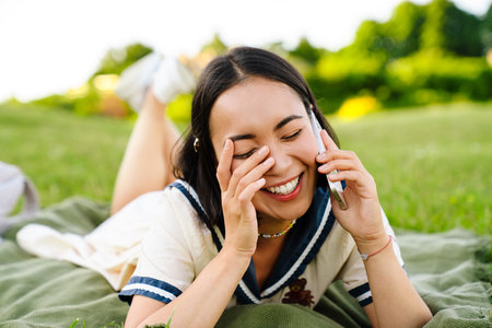 Cheerful asian woman wearing dress talking on mobile phone while lying on blanket in green parkの写真素材
