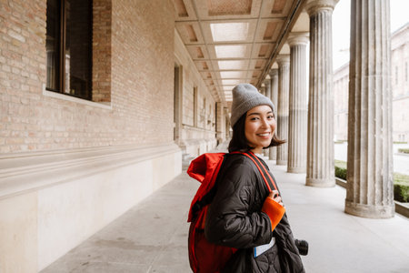 Beautiful smiling young asian woman tourist with backpack and camera walking through the old city streetの写真素材
