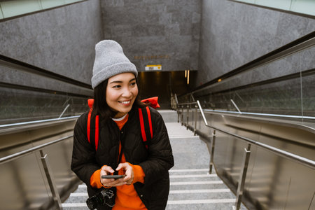 Beautiful young smiling asian woman in warm clothes using smartphone while climbing the stairs...