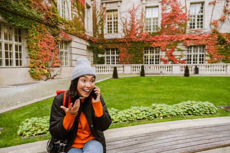 Surprised young asian woman in warm clothes talking on mobile phone while sitting on bench in old cityの写真素材