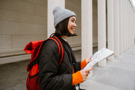 Young joyful asian woman in warm clothes examining paper map during walk at city streetの写真素材