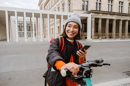 Young joyful asian woman in warm clothes using mobile phone for electric scooter's rent outdoors at city streetの写真素材