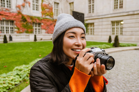 Beautiful young smiling asian woman in warm clothes taking pictures with vintage camera while sitting in old cityの写真素材