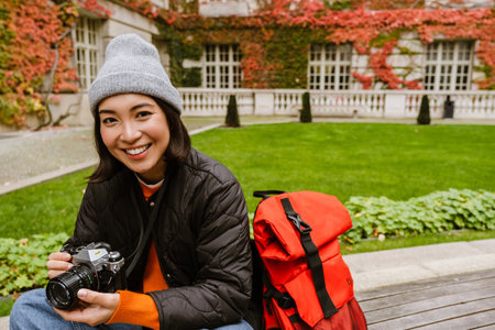 Beautiful young smiling asian woman in warm clothes taking pictures with vintage camera while sitting on bench in old cityの写真素材