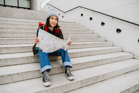 Young beautiful asian woman tourist in warm clothes examining paper map while sitting on stairs...