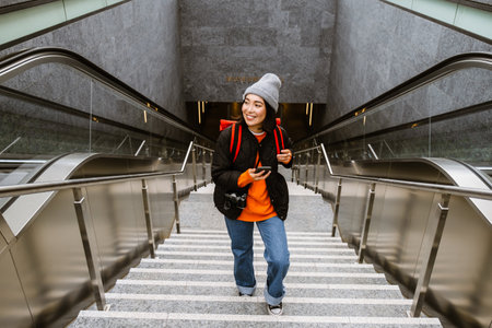 Beautiful young smiling asian woman in warm clothes using smartphone while climbing the stairs outdoorsの写真素材