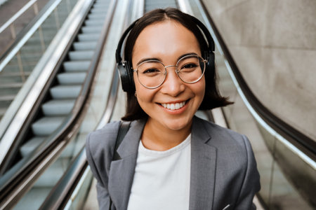 Young pretty asian woman in headphones smiling at camera while standing on escalator outdoorsの写真素材