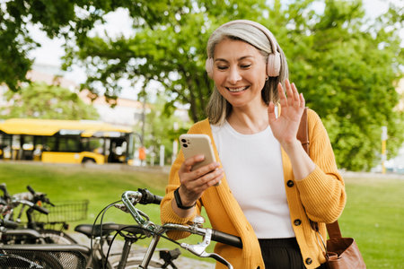 Grey asian woman gesturing and using cellphone while standing by her bicycle in park