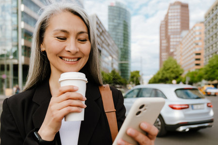 Grey asian woman drinking coffee and using mobile phone at city streetの写真素材