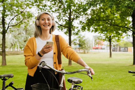 Grey asian woman laughing and using cellphone while standing by her bicycle in parkの写真素材