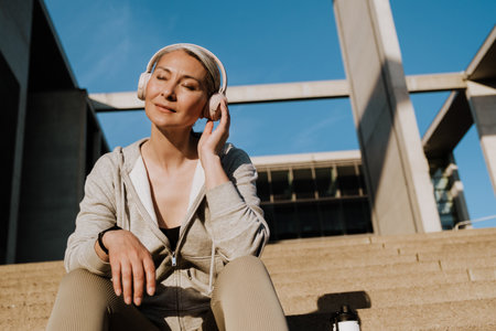 Asian mature woman in headphones listening to music and sitting on stairs during workout outdoors
