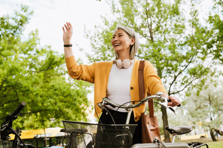 Grey asian woman laughing and gesturing while standing by her bicycle at summer park