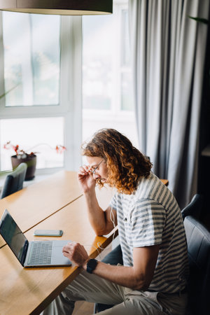 Young curly man using laptop and earphones while sitting by desk in officeの写真素材