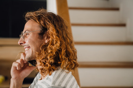 Young curly man smiling while talking on cellphone by stairs in officeの写真素材