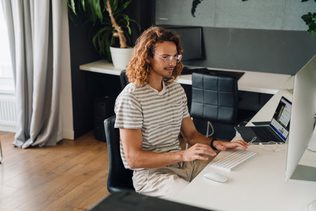 Young employee working on desktop computer and laptop while sitting at table in modern office