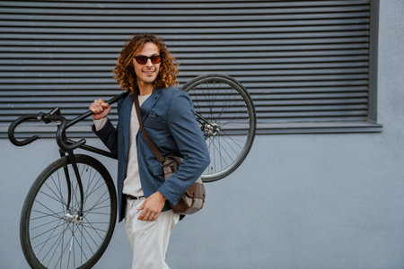 Ginger european man in sunglasses carrying bicycle near grey wall outdoors on city street