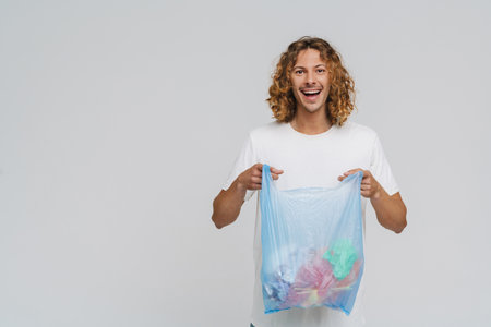 Ginger european man wearing t-shirt smiling while holding plastic bag with trash isolated over white...