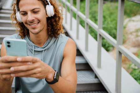 Portrait of young long-haired athletic smiling man in headphones using phone , while standing on the stairs outdoorsの写真素材