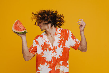 Ginger man in sunglasses laughing while dancing with watermelon isolated over yellow background