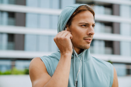 Young long-haired serious handsome man putting headphones in ear and looking aside , while standing near modern building outdoorsの写真素材