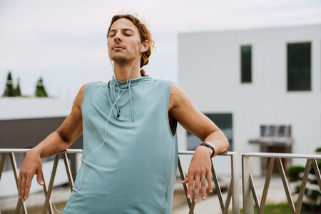 Young handsome athletic long-haired man in headphones relaxing with closed eyes, while leaning on...