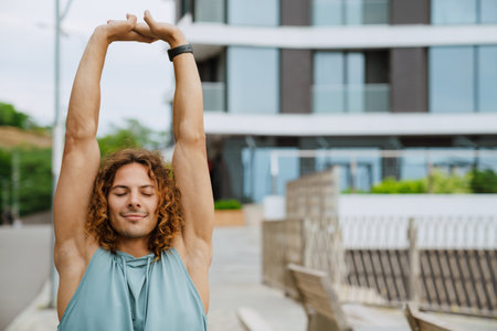 Young athletic long-haired handsome smiling man with closed eyes stretching his arms , while standing outdoorsの写真素材