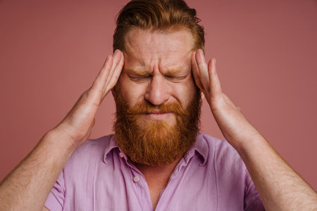 Portrait of adult tense redhead bearded man with closed eyes touching his temples, , while standing over isolated coral backgroundの写真素材