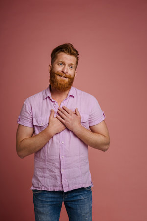 Adult handsome redhead bearded man with folded hands on chest expressing tender emotion and looking aside, while standing over isolated coral backgroundの写真素材
