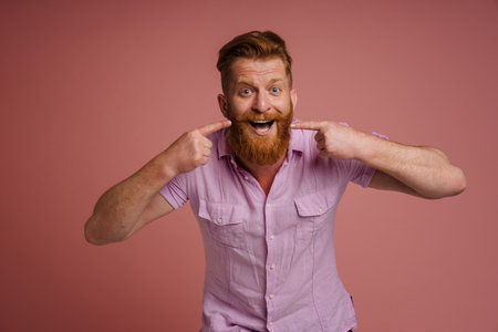 Ginger white man laughing and pointing fingers at his smile isolated over pink backgroundの写真素材