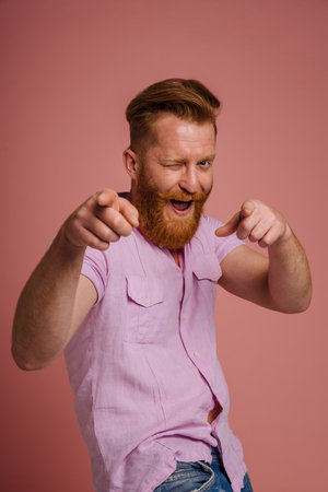 Ginger white man winking and pointing fingers at camera isolated over pink backgroundの写真素材