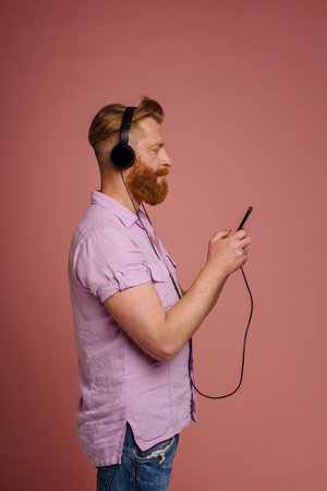 Adult stylish redhead bearded calm man in headphones with phone, standing with his side over...