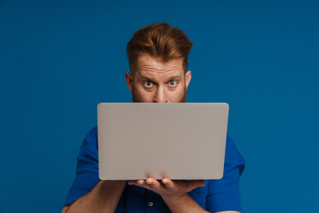 Ginger shocked man looking at camera while posing with laptop isolated over blue background