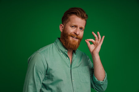 Ginger white man with beard gesturing and looking at camera isolated over green backgroundの写真素材