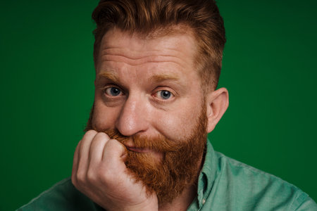 Portrait of adult handsome stylish bearded bored man propping his head and looking at camera , while standing over isolated green backgroundの写真素材