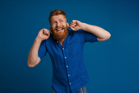 Ginger displeased man grimacing while plugging his ears isolated over blue backgroundの写真素材
