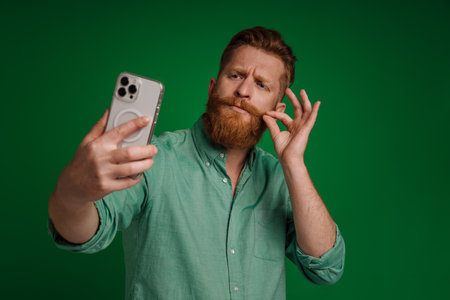 Ginger white man gesturing while taking selfie on mobile phone isolated over green backgroundの写真素材