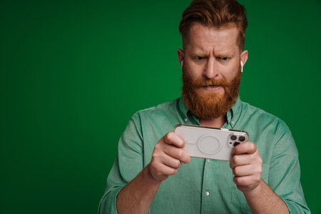 Ginger perplexed man in earphones playing online game on cellphone isolated over green background
