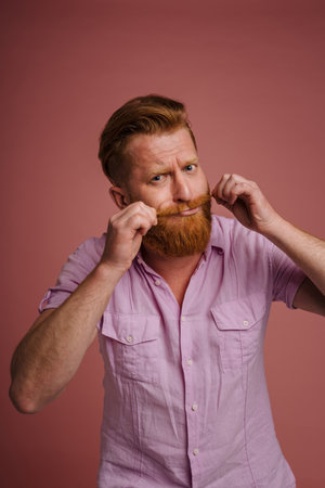 Ginger white man grimacing while making fun with mustache isolated over pink backgroundの写真素材