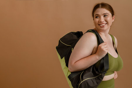 Young beautiful smiling plus size girl in training suit with bag looking aside , while standing over isolated brown backgroundの写真素材