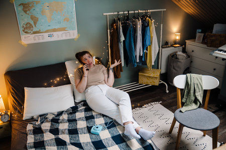 Young beautiful girl talking phone, gesturing and looking aside, while lying on bed in cozy room at homeの写真素材